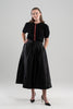 Woman wearing black puff-sleeve cardigan with red tartan plain ribbon trim and gold buttons, black skirt and black patent sling-back shoes on plain background.