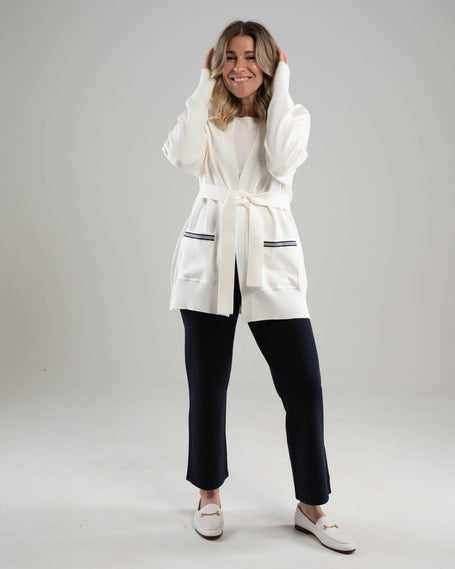 Woman wearing Noyac Knits ecoviscoe white long belted cardigan with navy and white striped ribbon trim on pockets, styled with Noyac Knits straight leg ankle length pants in navy and white loafers  on a plain background