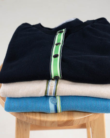 Stack of Noyac Knits cotton ribbon cardigans, one in sky blue, one in light tan, one in navy folded on a wooden stool with a plain background