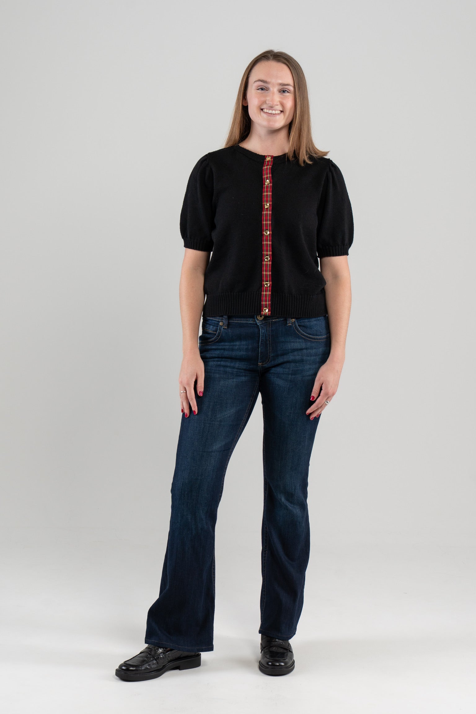 Woman wearing black puff-sleeve cardigan with red tartan plaid ribbon trim and gold buttons, jeans, loafers on plain background.