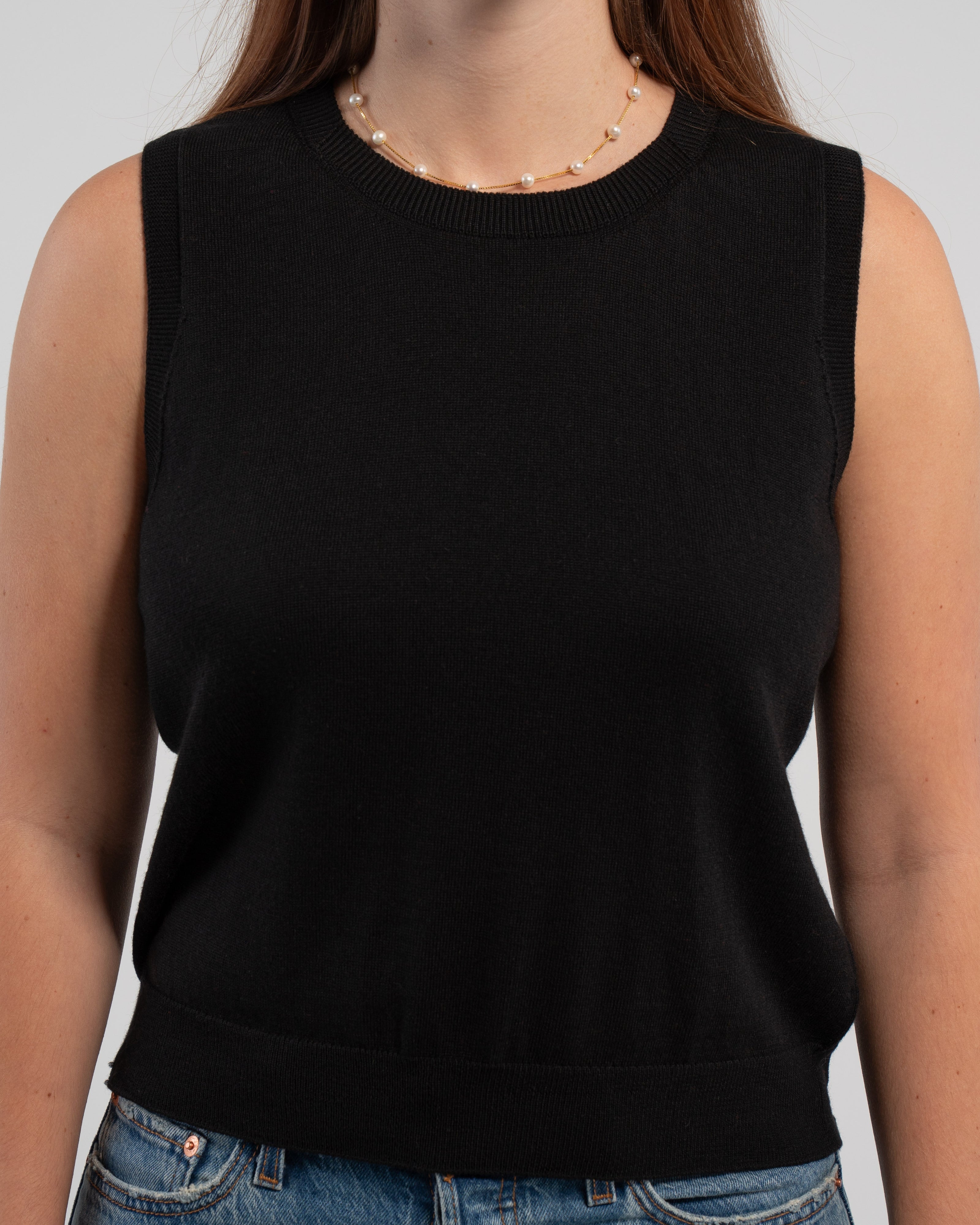 Close up of woman wearing Noyac Knits classic sleeveless sweater shell in black knit in ultra soft premium lightweight merino styled with jeans, pearl choker on plain background.  This versatile piece works well alone, over a blouse, under a matching classic cardigan or under a blazer.  