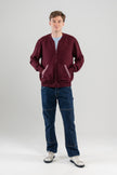 Man wearing Noyac Knits bomber-style sweater in burgundy color with signature striped ribbon-trimmed pockets -- knit in premium, ultrasoft merino wool -- styoed with  light blue t-shirt, jeans and sneakers on plain background.