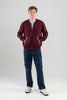 Man wearing Noyac Knits bomber-style sweater in burgundy color with signature striped ribbon-trimmed pockets -- knit in premium, ultrasoft merino wool -- styoed with  light blue t-shirt, jeans and sneakers on plain background.