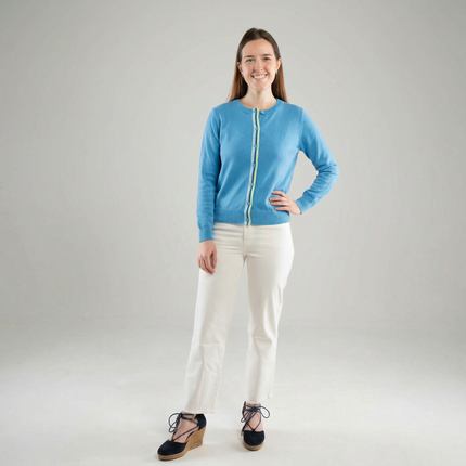 Woman wearing a Noyac Knits the Allison sky blue cardigan in breathable, premium cotton and trimmed with beautiful green and blue striped ribbon styled with  white pants and summer espadrilles on white background