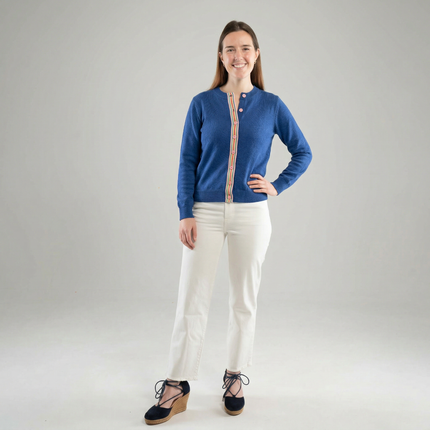 Woman wearing Noyac Knits cobalt blue classic length cardigan with signature pink rainbow ribbon trim and pink buttons, made in premium lightweight merino, styled with white jeans and blue espadrilles on plain background