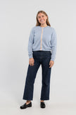 Woman wearing buttoned Noyac Knits Mary crop cardigan in soft, premium merino and trimmed with white mother of pearl buttons and signature ribbon in stripes of pastel pink, blue and green striped grosgrain ribbon styled with jeans and loafers on plain background.