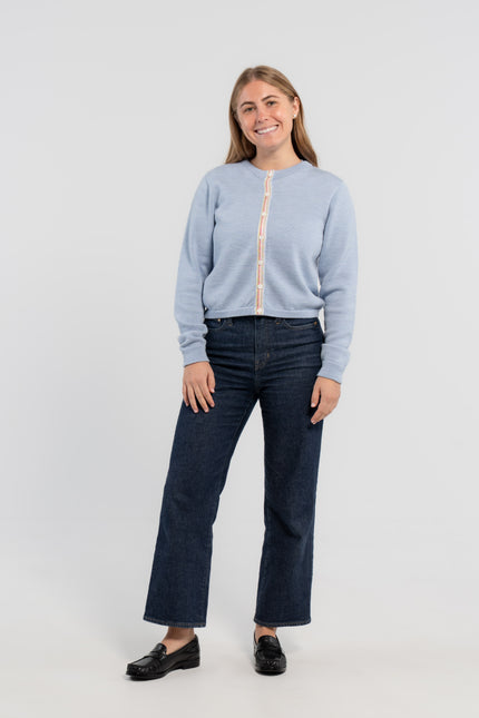 Woman wearing buttoned Noyac Knits Mary crop cardigan in soft, premium merino and trimmed with white mother of pearl buttons and signature ribbon in stripes of pastel pink, blue and green striped grosgrain ribbon styled with jeans and loafers on plain background.