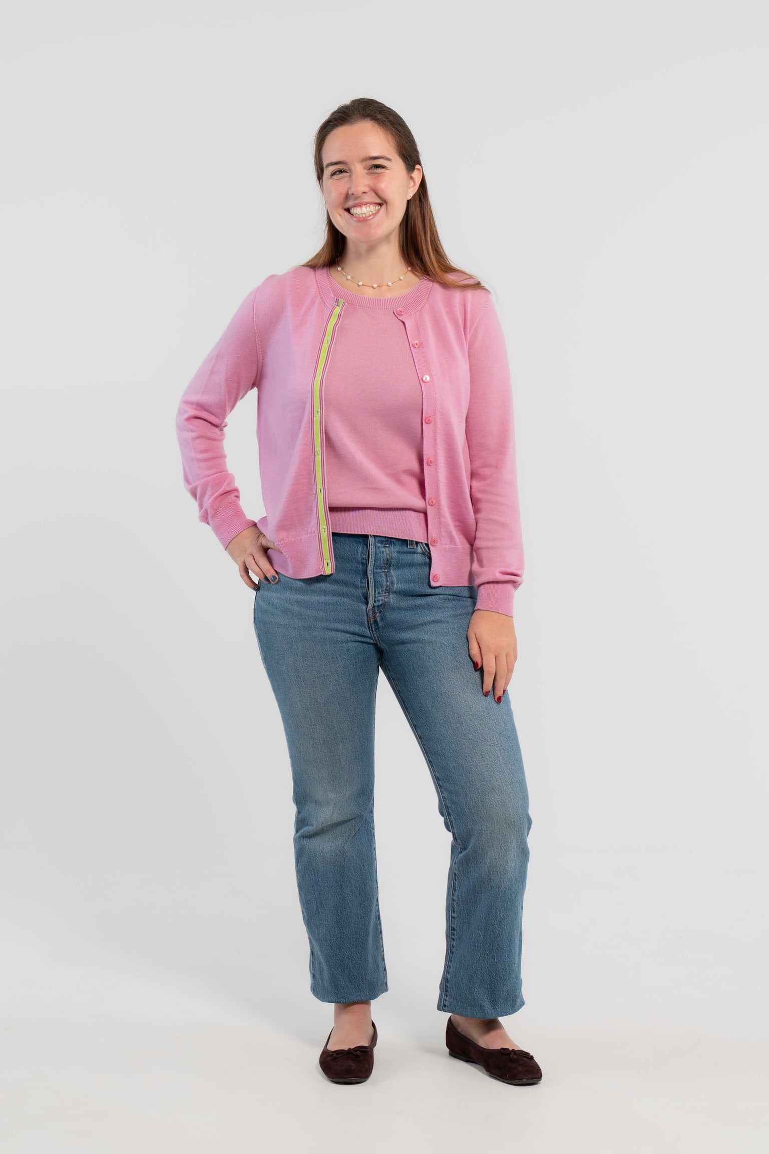 Picture of woman wearing Noyac Knits orchid pink cardigan with pink buttons and  signature ribbon trim in stripes of green, pink and lilac -- knit in ultrasoft premium merino wool, styled with pearl choker necklace, jeans, and ballet flats on plain bakckground.