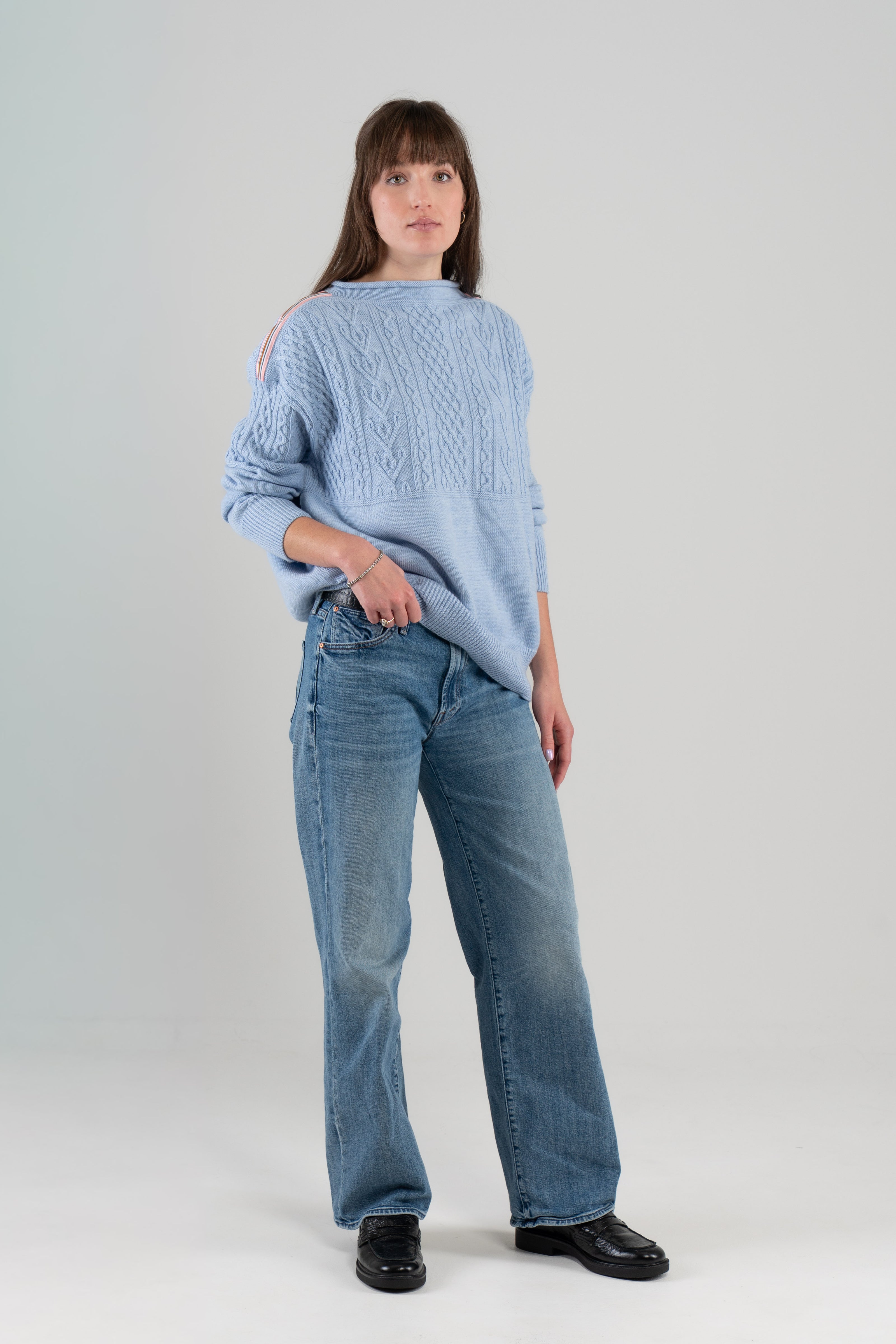 Woman wearing Noyac Knits Channing boatneck pullover in beautiful baby blue featuring cabling and signature ribbon trim on shoulder, knit in premium soft merino wool and styled with  jeans and loafers on plain background.
