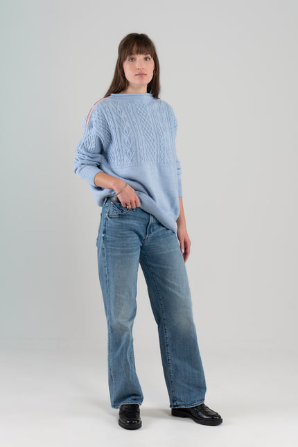 Woman wearing Noyac Knits Channing boatneck pullover in beautiful baby blue featuring cabling and signature ribbon trim on shoulder, knit in premium soft merino wool and styled with  jeans and loafers on plain background.