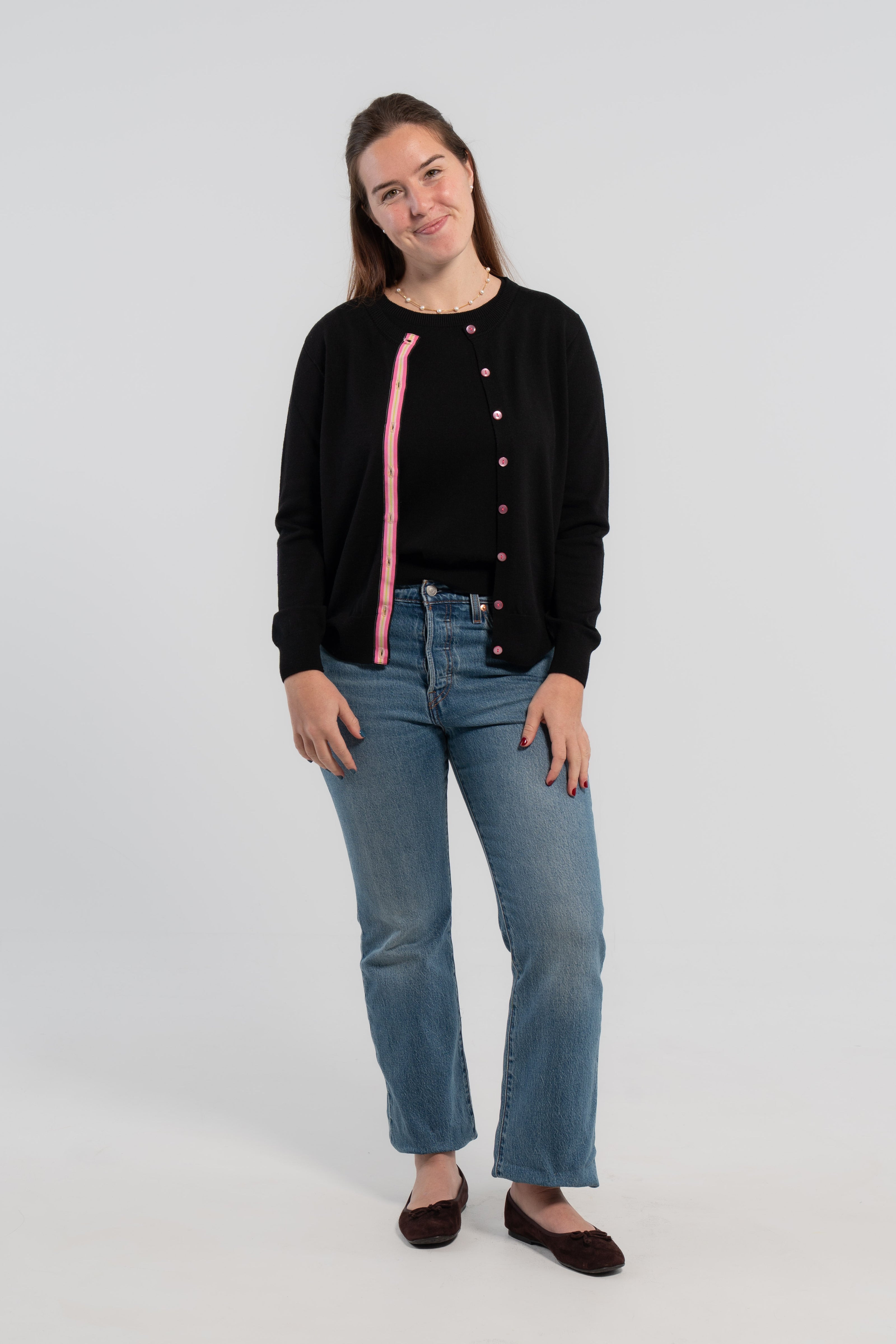Woman wearing Noyac Knits Kate Classic Cardigan in black premium lightweight merino and trimmed with signature pink buttons and hot pink, light pink and green striped grosgrain ribbon trim styled over matching black merino crew neck sleeveless sweater shell, jeans, ballet flats on plain background.
