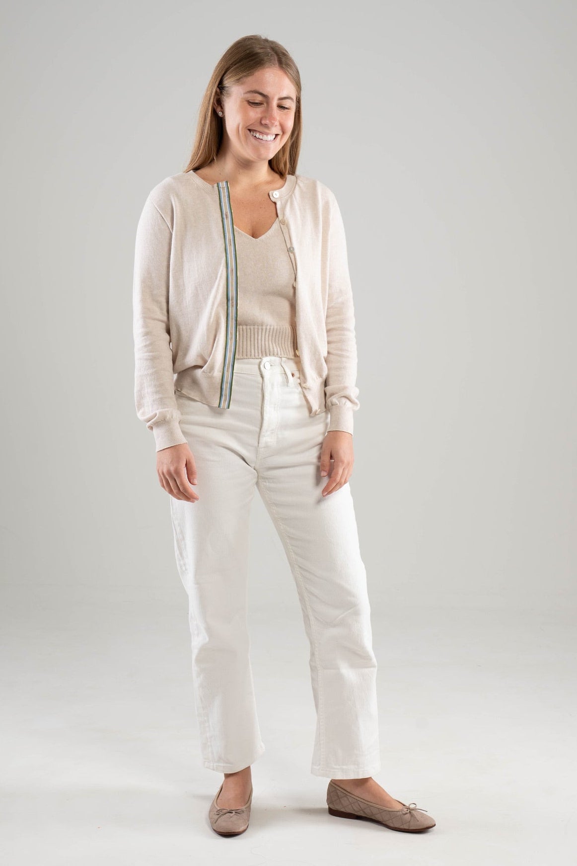 Woman wearing Noyac Knits Lynn classic cardigan in light tan  with army green, light blue, white, and tan striped ribbon trim and mother of pearl buttons, in premium Egyptian cotton, styled with matching Noyac Knits double v-neck cotton tank and white jeans and ballet flats on  white background 