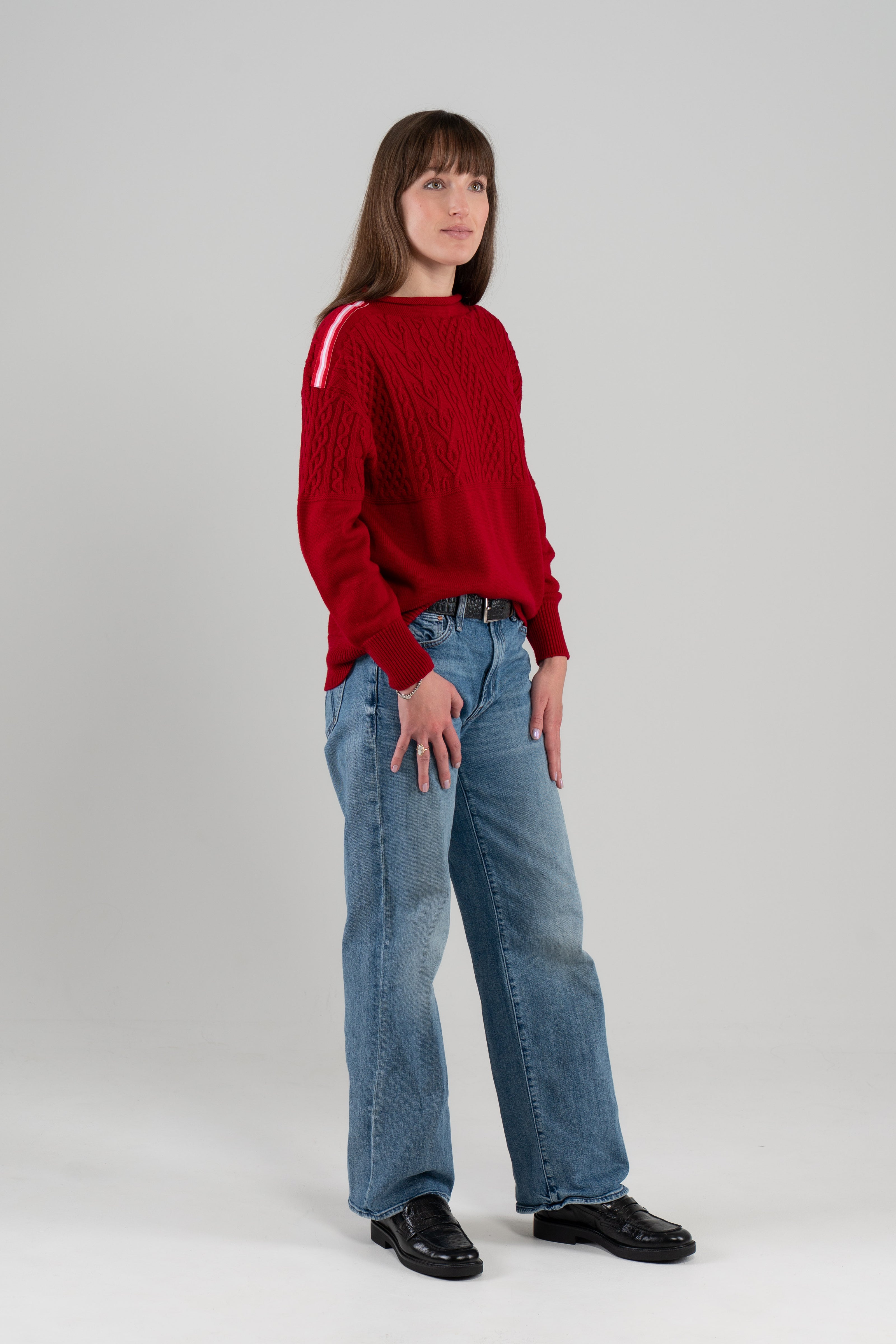 Woman wearing Noyac Knits Rhoda boatneck pullover sweater with beautiful cabling and signature red, pink and white striped ribbon trim on shoulders, knit in ultra soft premium merino wool, and styled with jeans and loafers on plain background.