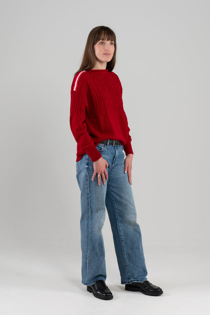Woman wearing Noyac Knits Rhoda boatneck pullover sweater with beautiful cabling and signature red, pink and white striped ribbon trim on shoulders, knit in ultra soft premium merino wool, and styled with jeans and loafers on plain background.