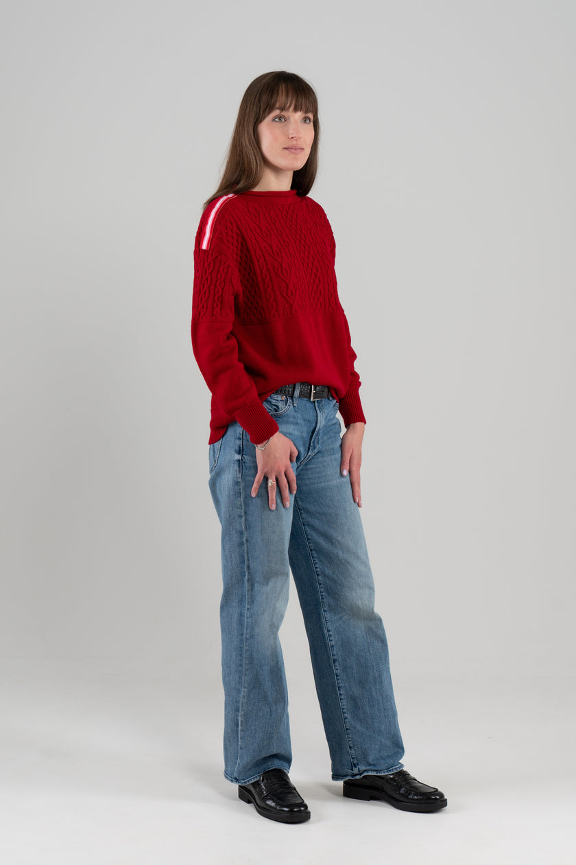 Woman wearing Noyac Knits Rhoda boatneck pullover sweater with beautiful cabling and signature red, pink and white striped ribbon trim on shoulders, knit in ultra soft premium merino wool, and styled with jeans and loafers on plain background.