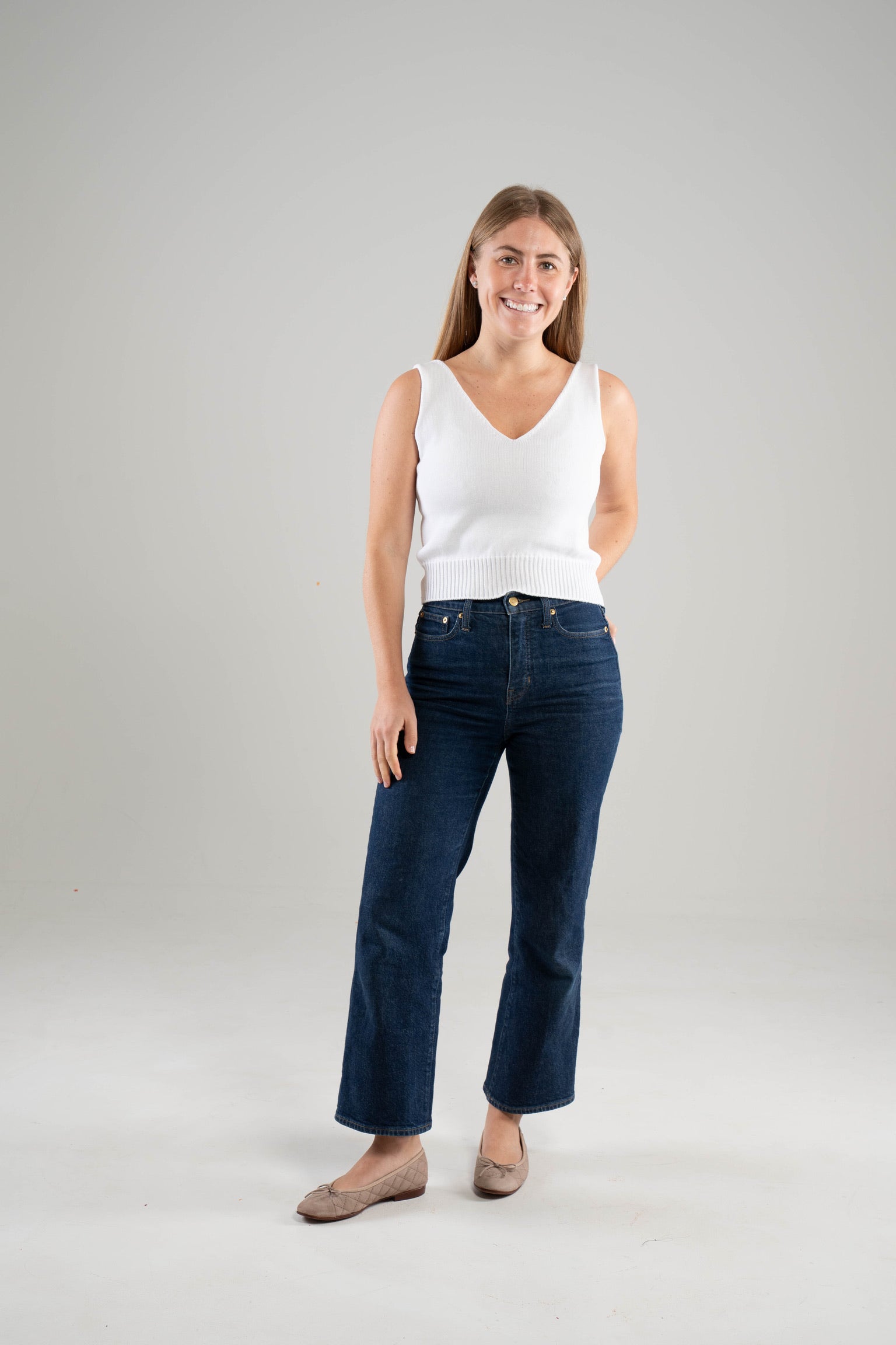 Woman wearing a Noyac Knits white cotton double v-neck crop tank top styled with dark blue jeans and ballet flats on a white  background