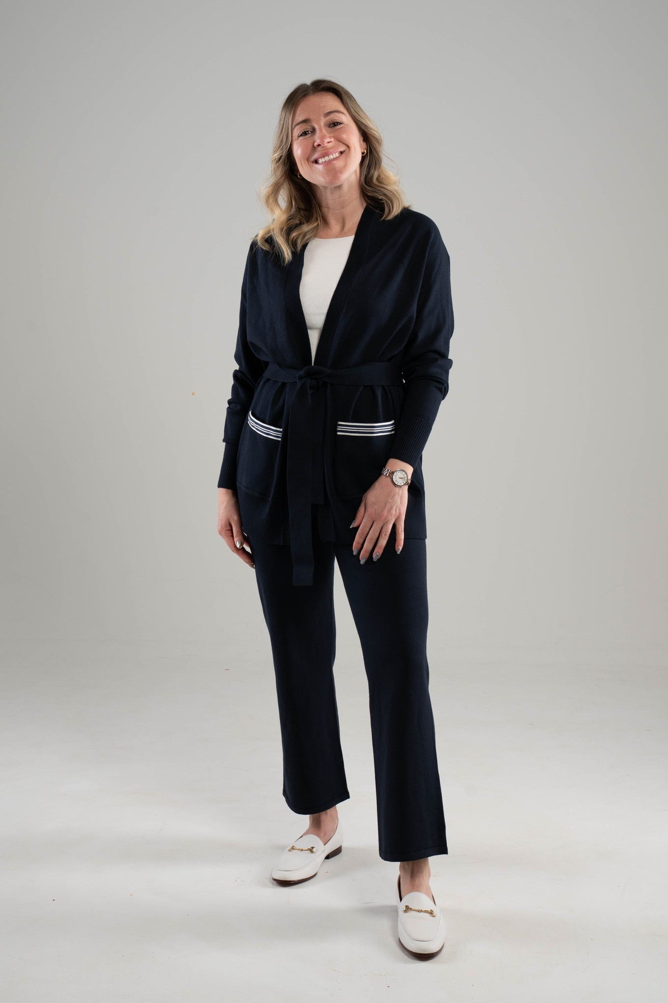 Woman wearing a Noyac Knits long belted navy cardigan with navy and white ribbon-trimmed pockets over white featherweight scoop neck sleeveless sweater and navy knit ankle length pants -- all in environmentally sustainable ecoviscose on a white background