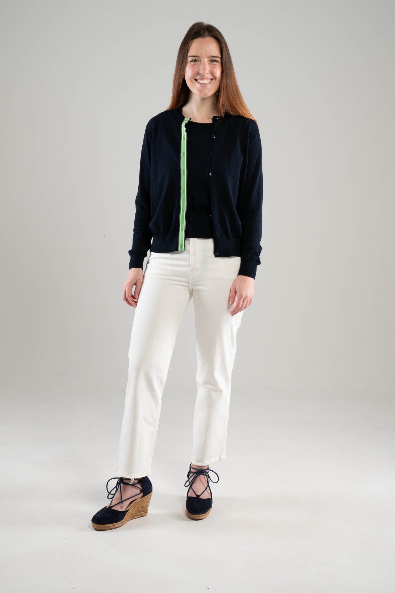 Woman wearing Noyac Knits navy Julie ribbon cardigan trimmed with with green, navy and white striped ribbon over a navy crew neck sleeveless sweater, both in premium Egyptian cotton, styled with white jeans and navy espadrilles  on a white background