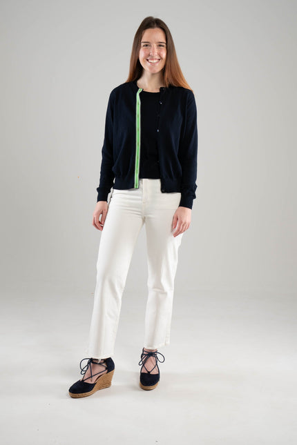 Woman wearing Noyac Knits navy Julie ribbon cardigan trimmed with with green, navy and white striped ribbon over a navy crew neck sleeveless sweater, both in premium Egyptian cotton, styled with white jeans and navy espadrilles  on a white background