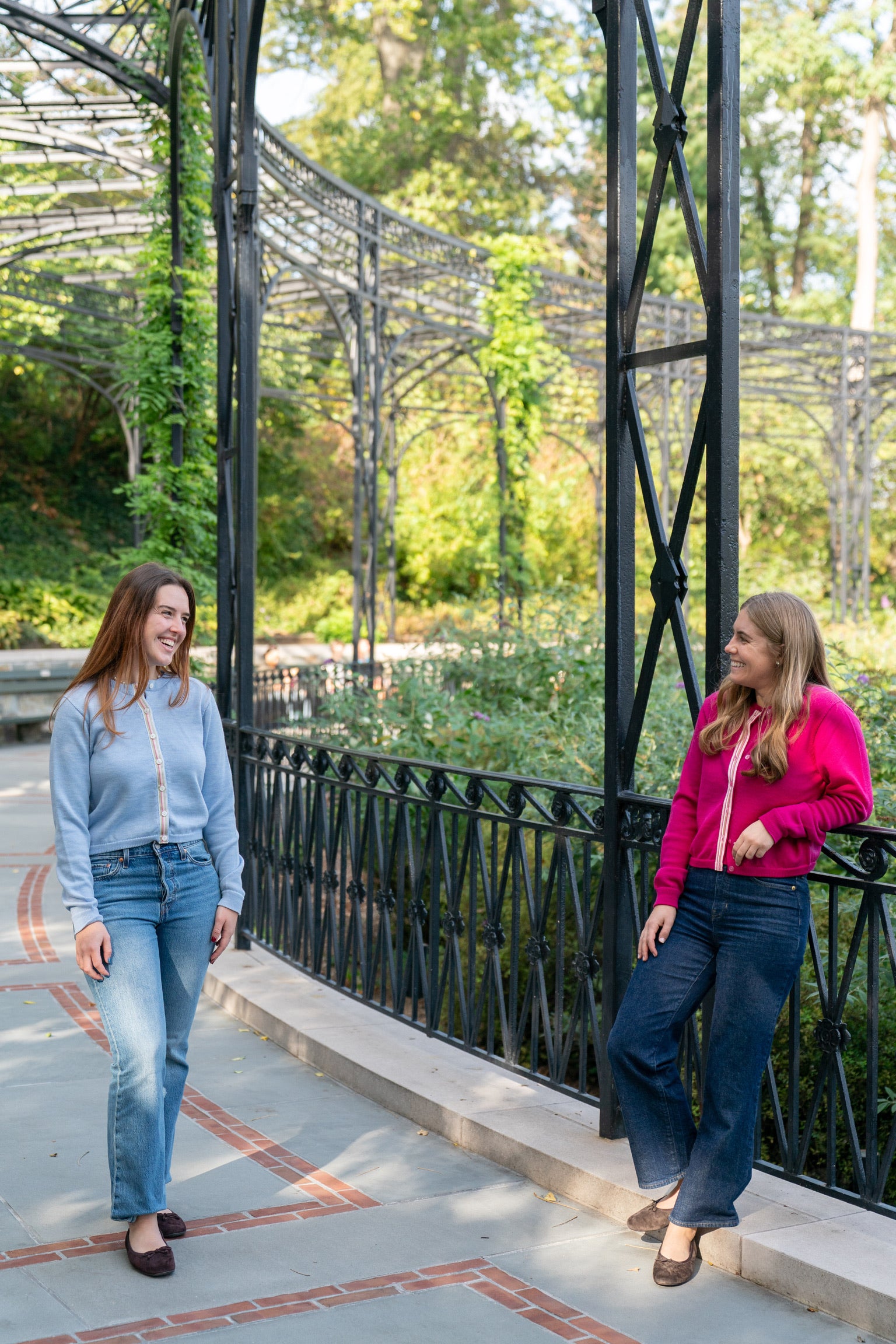 Two women walking and talking on a paved path with a decorative metal archway in the background wearing colorful Noyac Knits crop cardigans knit in premium merino wool styles with jeans and ballet flats.
