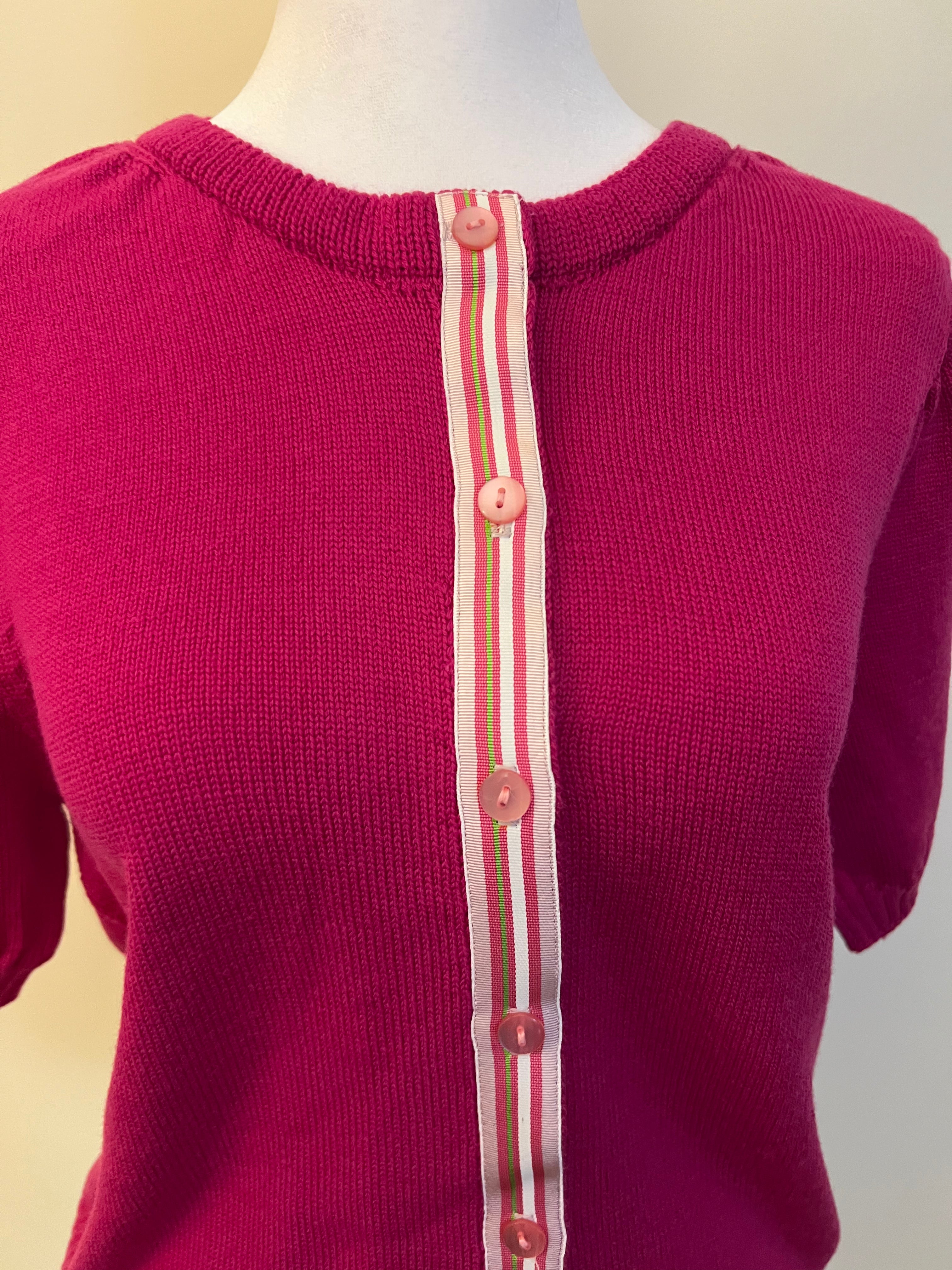 Raspberry pink sweater with a striped ribbon trim in light and dark pink, white and green stripes and pink buttons on a mannequin against a beige background