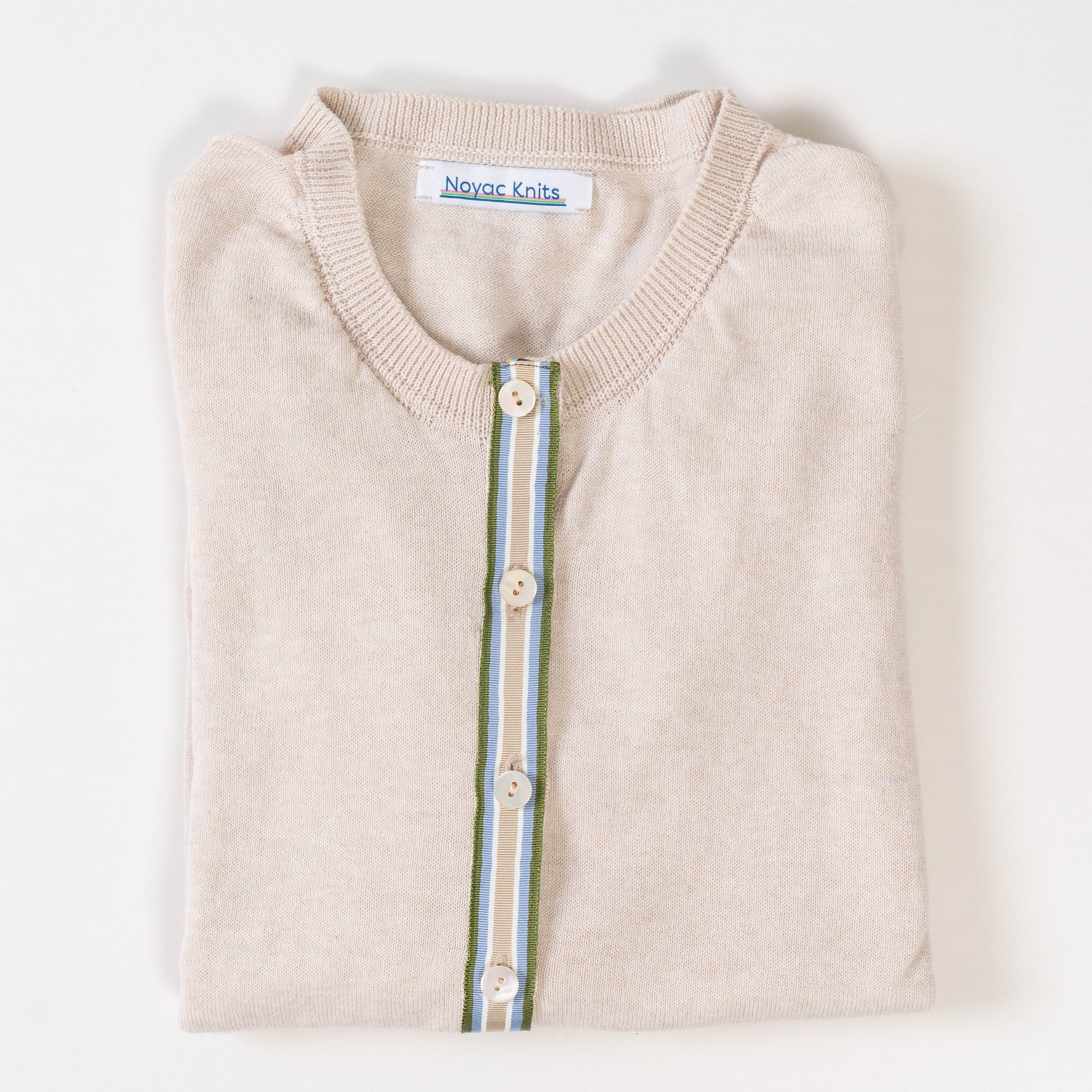 Close-up of Noyac Knits light tan cardigan with a army green, light blue, white, and tan striped ribbon trim and shell buttons over v-neck tan shell sweater on white background