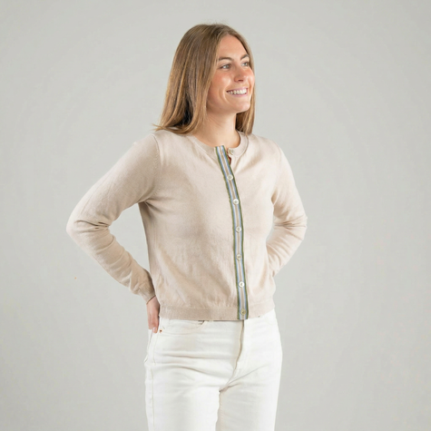 Woman wearing a Noyac Knits light tan ribbon cardigan with army green, light blue, white , and tan striped ribbon trim and shell butons, white jeans and white background 