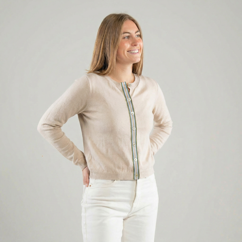 Woman wearing a Noyac Knits light tan ribbon cardigan with army green, light blue, white , and tan striped ribbon trim and shell butons, white jeans and white background 