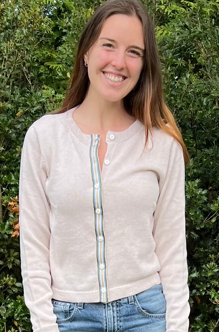 Woman wearing Noyac Knits sand colored crop cardigan in cotton trimmed with green, blue, tan and white striped ribbon and mother of pearl buttons in outdoor setting.