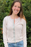 Woman wearing Noyac Knits sand colored crop cardigan in cotton trimmed with green, blue, tan and white striped ribbon and mother of pearl buttons in outdoor setting.