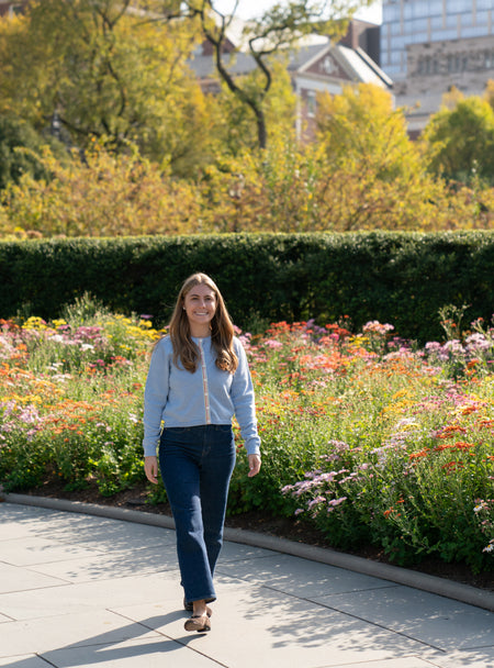 Woman wearing Noyac Knits Mary light blue crop cardigan with signature striped ribbon trim - knit in ultrafine merino wool - styled with jeans walking on a sidewalk with a garden and trees in the background.