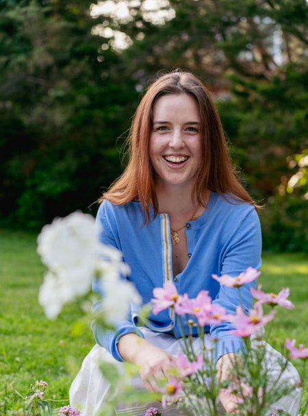 Woman in french blue cardigan with ribbon trim sitting in front of flowers with grass and trees in background