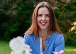 Woman in french blue cardigan with ribbon trim sitting in front of flowers with grass and trees in background