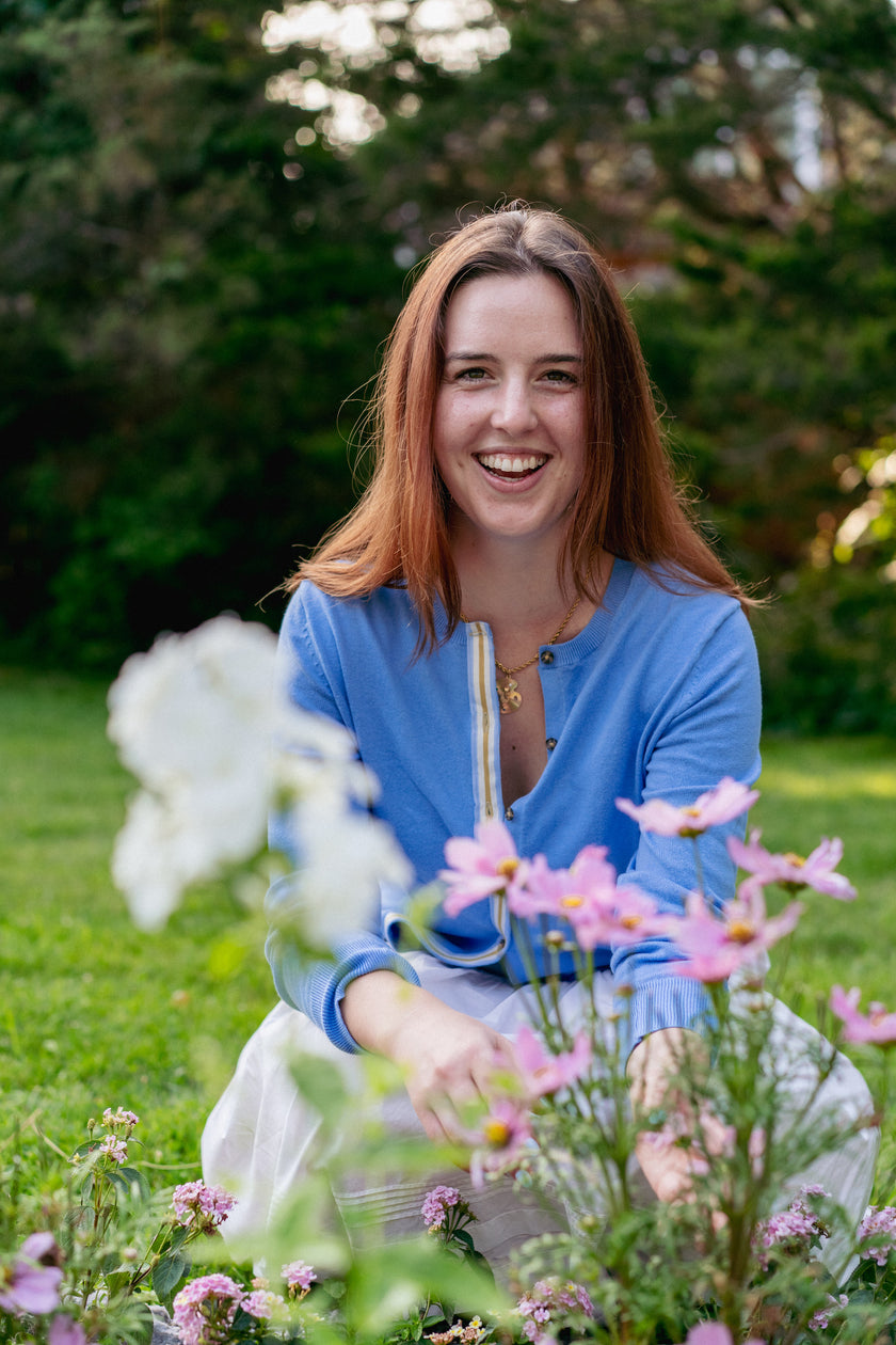 Woman in french blue cardigan with ribbon trim sitting in front of flowers with grass and trees in background