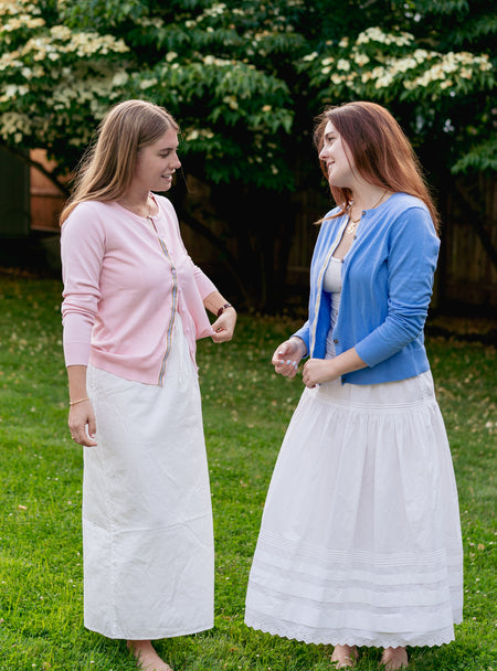 Two women wearing white cotton dresses and cardigans with grass and trees in background.  One cardigan is pink and one is french blue and both have ribbon trim.