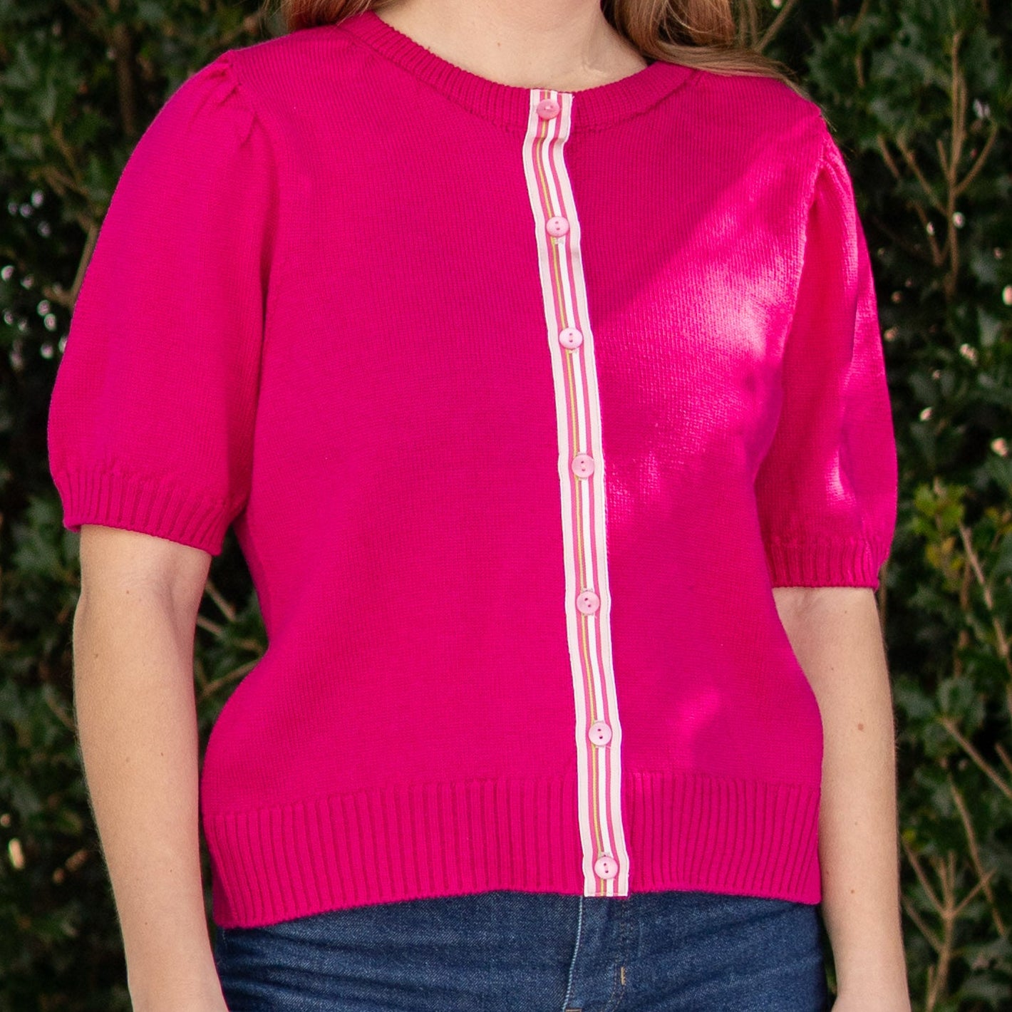 Woman wearing a dark pink cardigan with pink, white, green striped ribbon trim,  blue jeans standing outdoors with greenery in the background