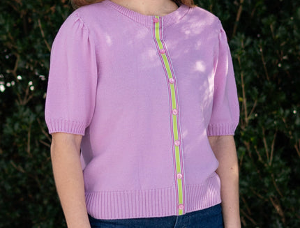 Woman wearing lilac pink short sleeve cardigan with pink and green ribbon trim, jeans against green leafy background