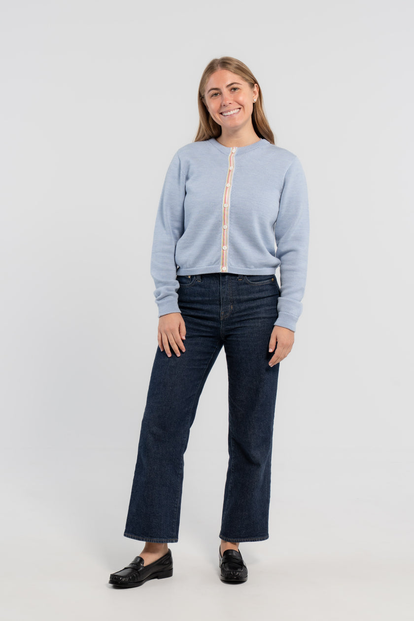 Woman wearing buttoned baby blue crop cardigan with white mother of pearl buttons and pastel pink, blue and green striped grosgrain ribbon trim, jeans, loafers on plain background.