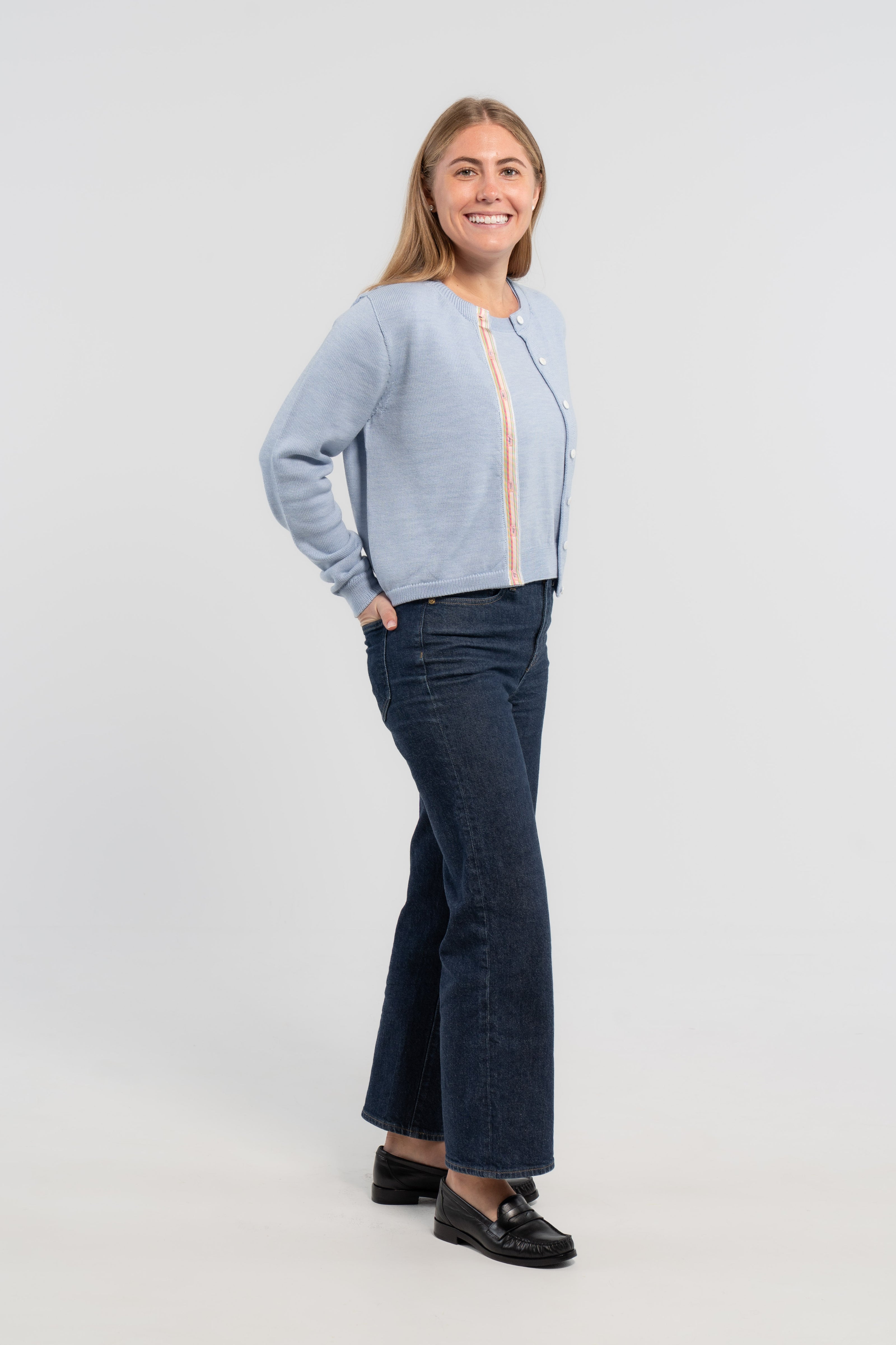 Women standing partially sideways on plain background wearing baby blue cardigan with pastel blue, pink and green striped grosgrain ribbon trim and white mother of pearl buttons, jeans, loafers.