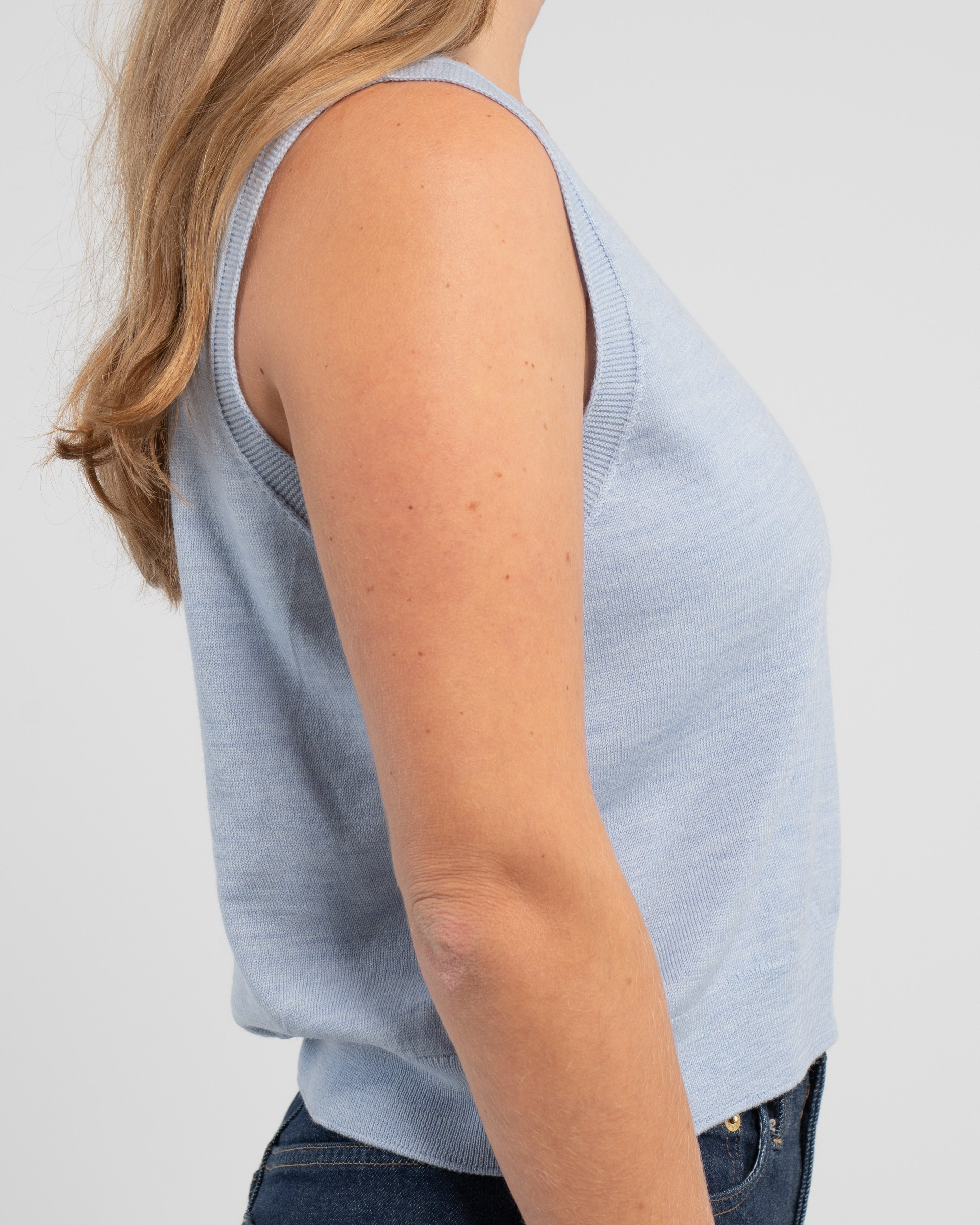 Sideview of woman wearing light blue crop sleeveless sweater and jeans on plain background