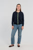 Woman wearing buttoned navy cardigan with navy buttons and green, white and navy striped grosgrain ribbon trim, jeans, ballet flats on plain background.