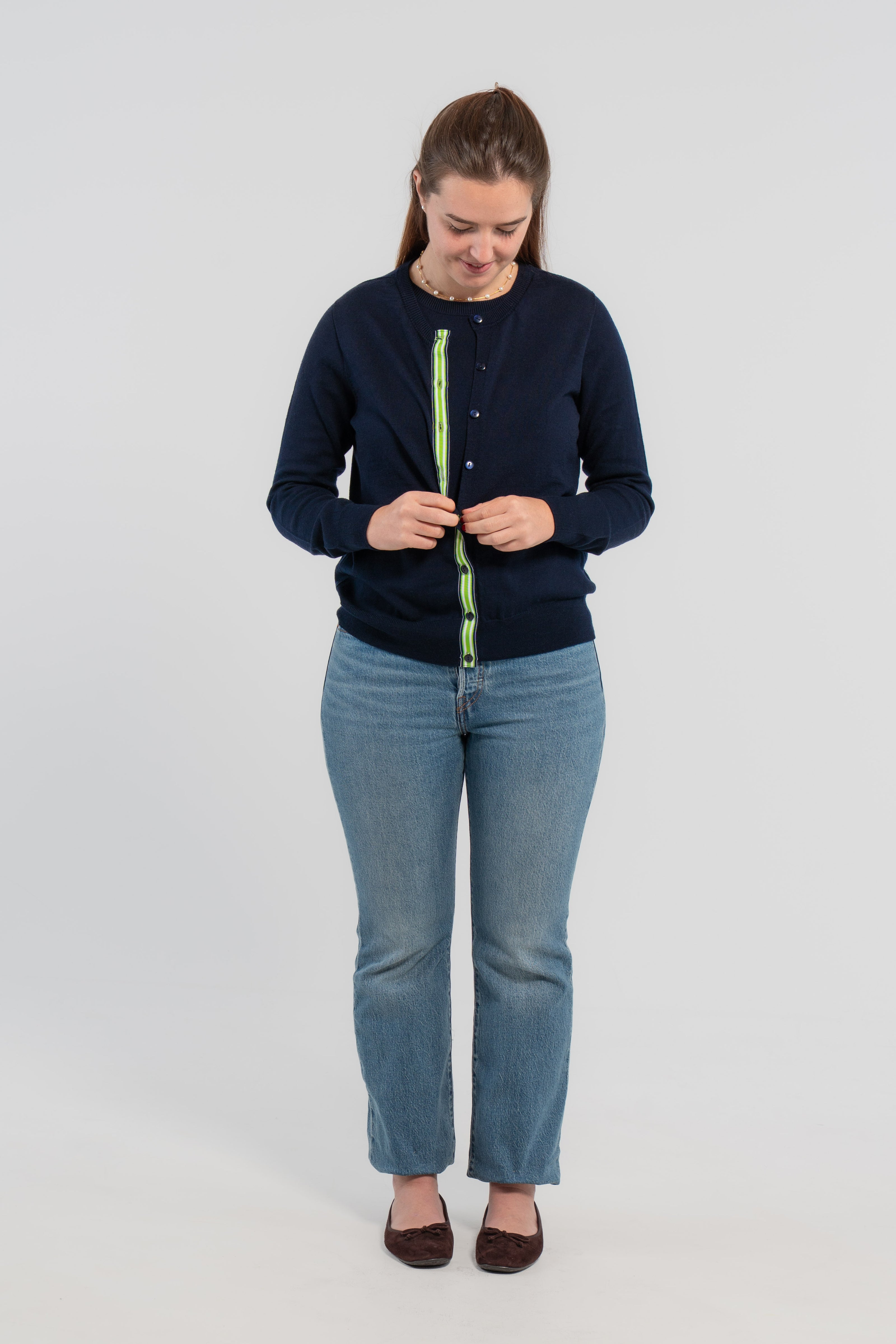 Woman buttoning while wearing a navy cardigan with navy buttons and navy, green and white striped ribbon trim, wearing jeans and ballet flats on plain background.