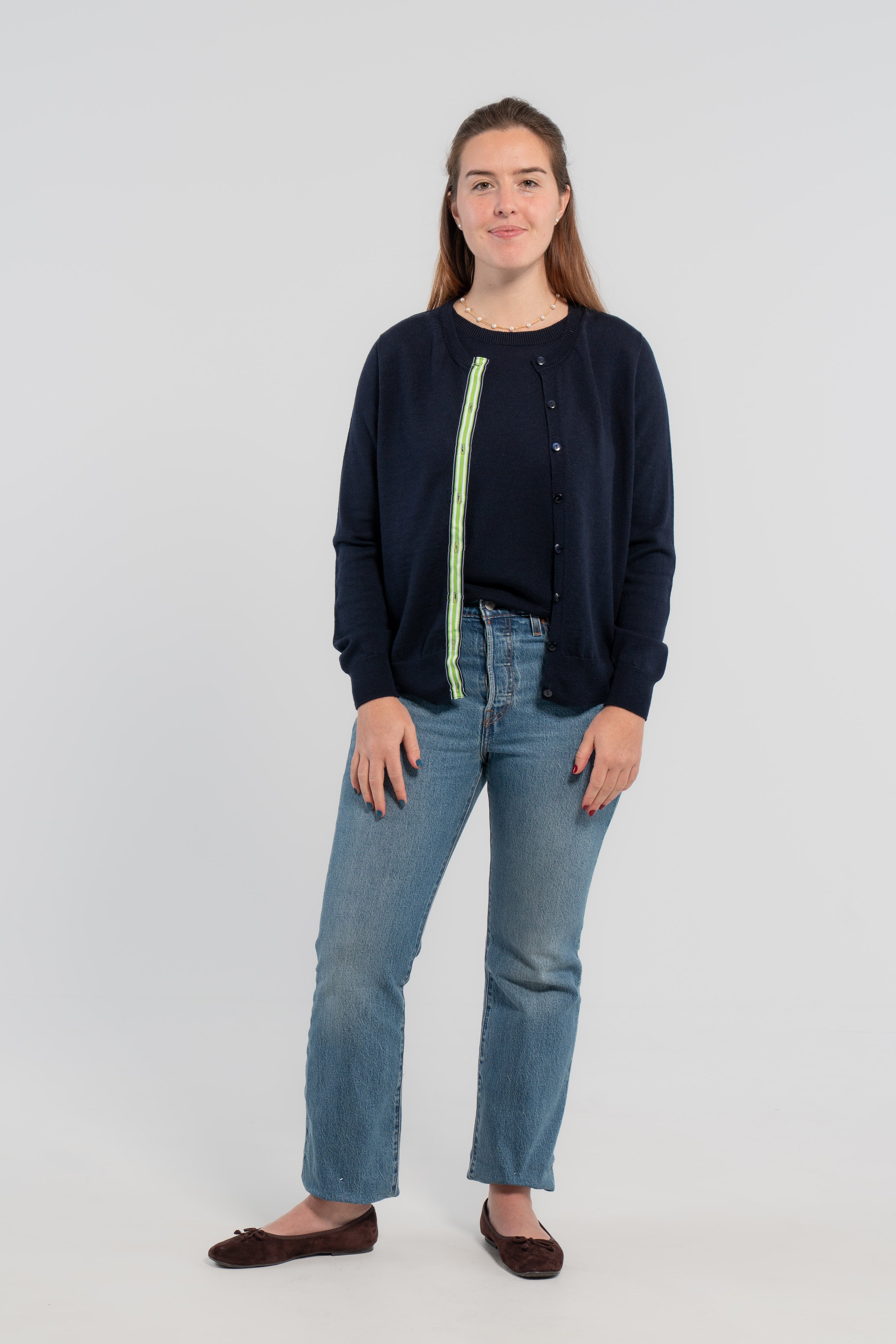 Woman wearing unbuttoned navy cardigan with navy buttons and green, white and navy grosgrain ribbon trim over matching navy shell sweater, jeans, ballet flats on plain background.