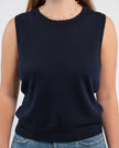 Close up of woman wearing navy sleeveless sweater, jeans, pearl choker neckless on plain background.