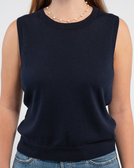 Close up of woman wearing navy sleeveless sweater, jeans, pearl choker neckless on plain background.