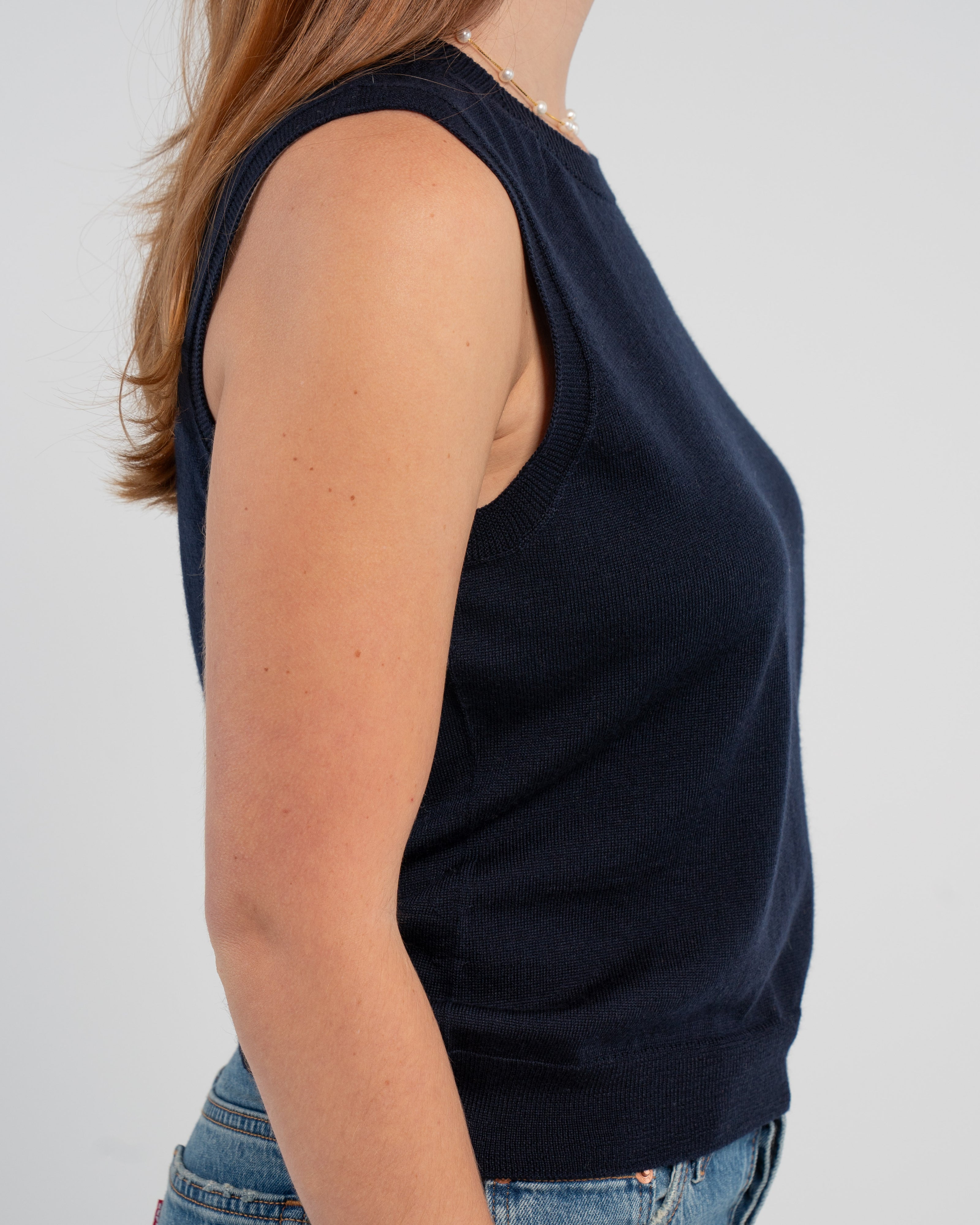 Sideview of woman wearing navy sleeveless sweater, jeans, pearl choker neckless on plain background.