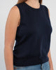 Close up of woman wearing navy sleeveless sweater, jeans, pearl choker neckless on plain background.
