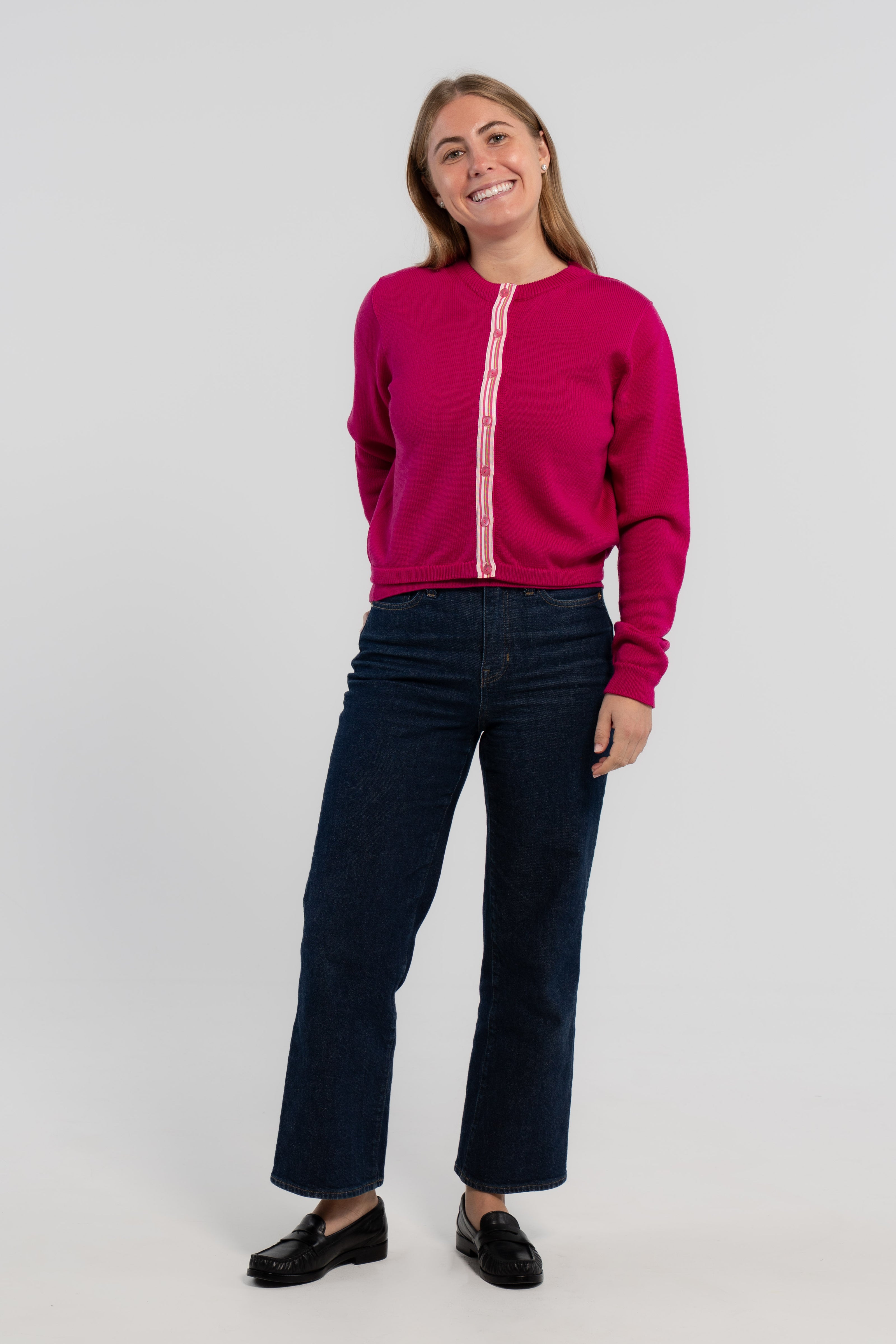 Woman wearing buttoned raspberry pink crop cardigan with pink buttons and pink, white, green striped grosgrain ribbon trim, jeans, loafers on plain background.