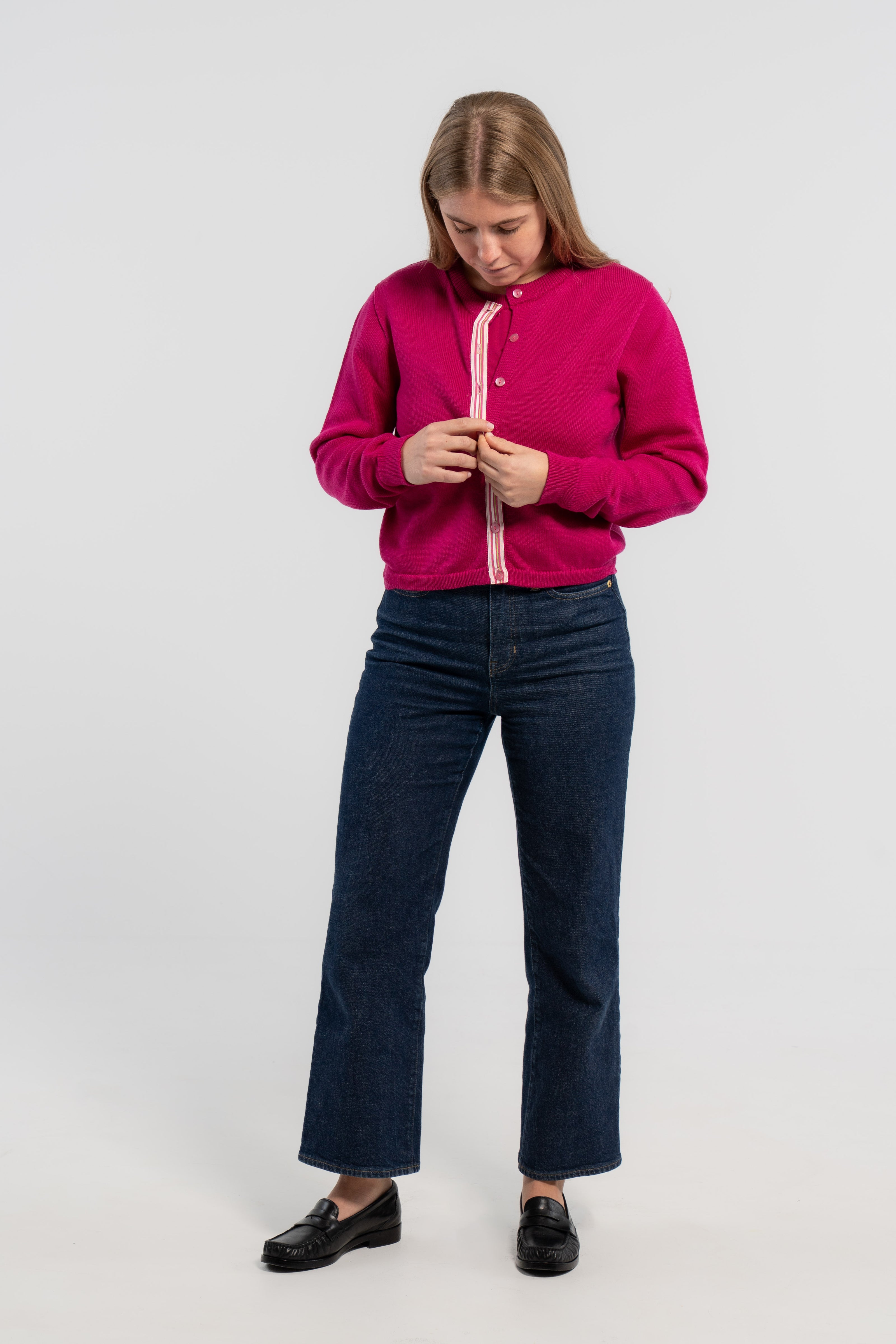 Woman buttoning raspberry pink crop cardigan with pink buttons and pink, white and green striped grosgrain ribbon trim, jeans, loafers on plain background.