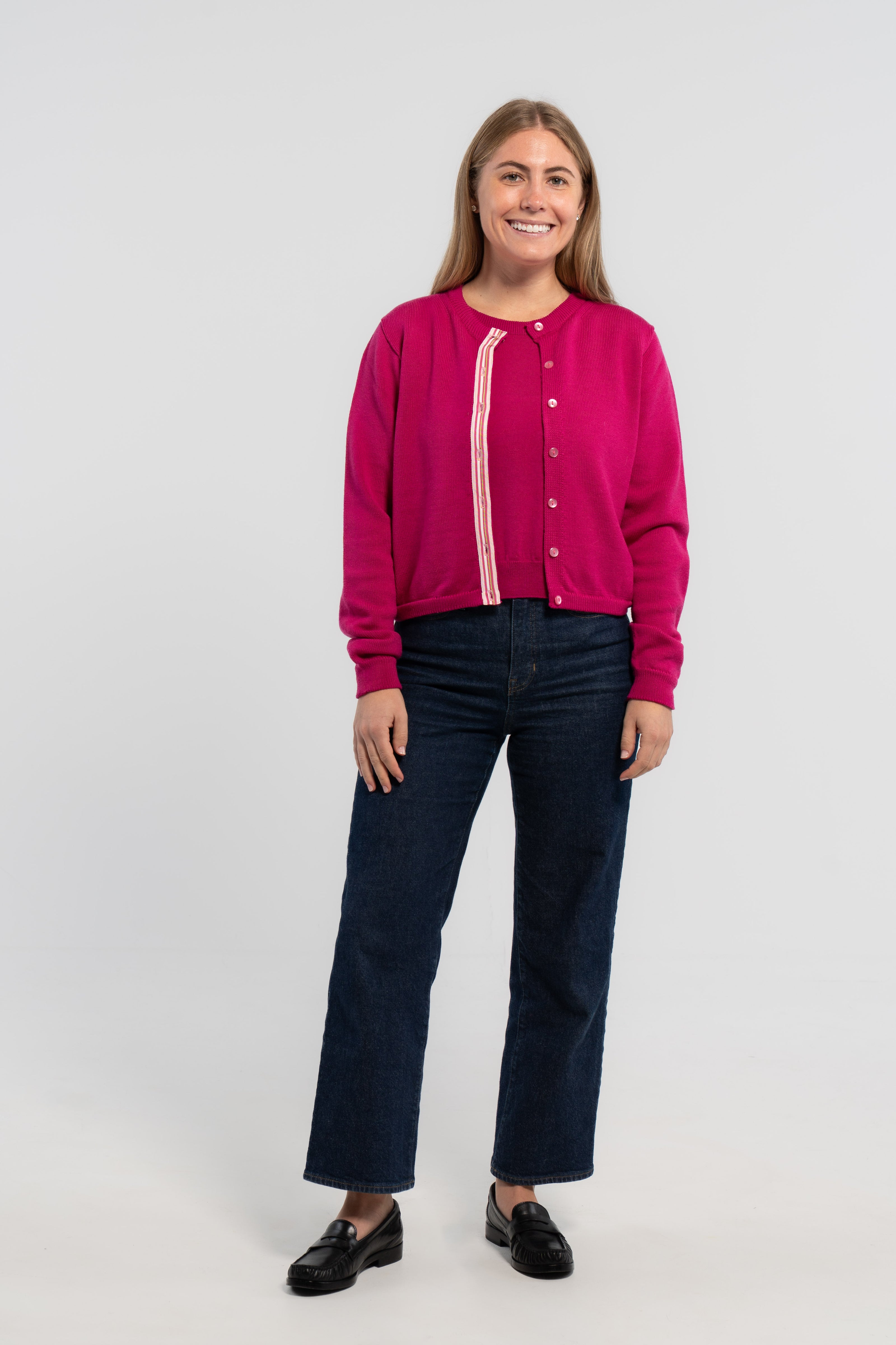 Woman wearing raspberry pink crop cardigan with pink buttons and pink, white and green striped grosgrain ribbon trim over matching raspberry pink sweater shell, jeans, loafers on plain background.